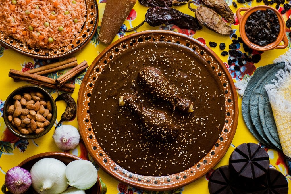 An overhead view of Oaxacan delicacies: Chicken in a dark brown mole sauce topped with sesame seeds, surrounded by chiles, rice, onions, and other spices.