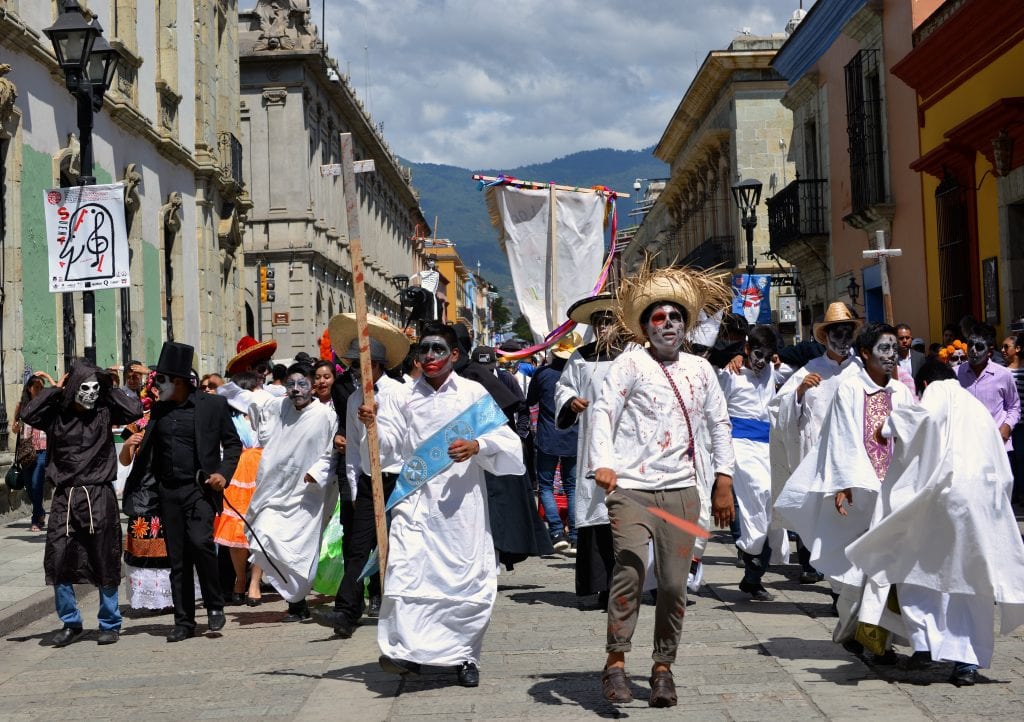 Oaxacan people dressed up in white clothing with zombie-like gray face makeup, parading through the streets for Day of the Dead.