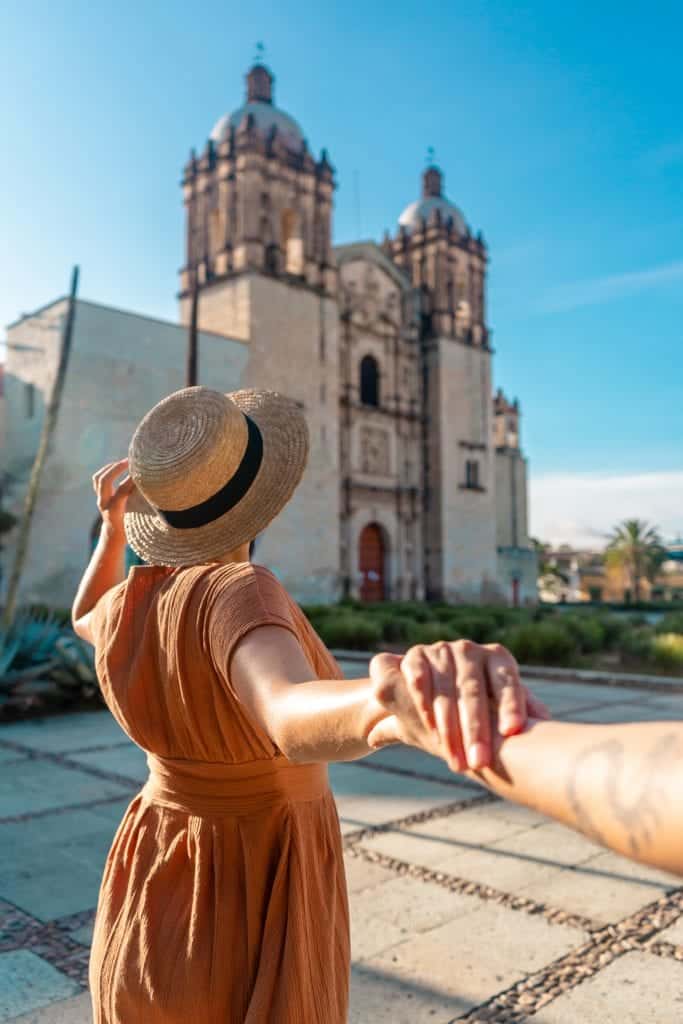 A woman posing for Instagram in front of Templo Santo Domingo in a wide-brimmed hat, holding the hand of the photographer behind her.