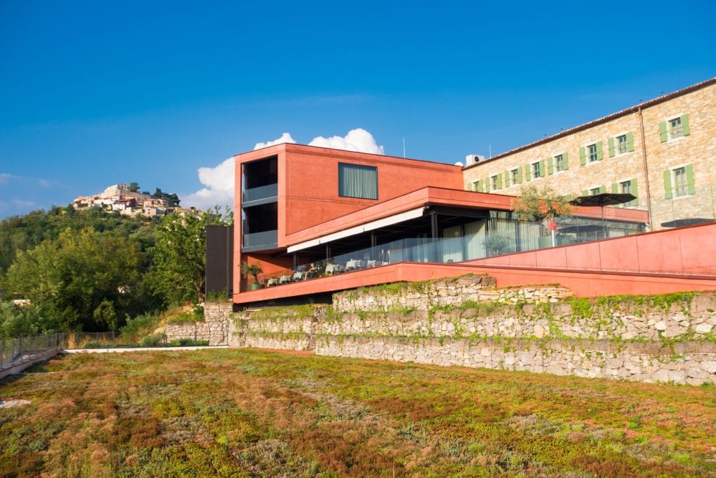 ROXANICH Wine Hotel, a modern pale red building with the Mountain View of Motovun in the background. In the foreground is a roof covered with green vegetation.