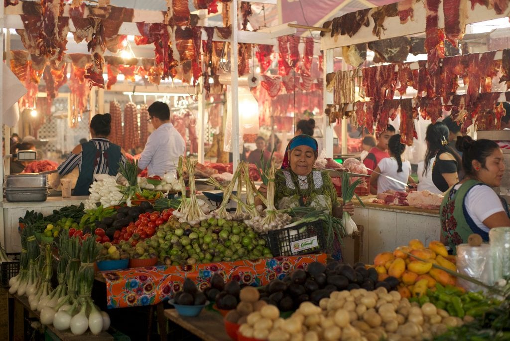 Indigenous women working in a market. There are tables full of fresh produce (limes, peppers, fennel) and raw meat hanging above.