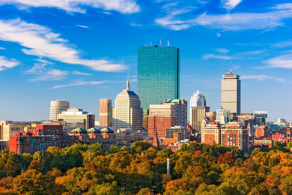 The skyline of Boston underneath a bright blue sky; at the bottom of the buildings are trees turning shades of orange, red, and yellow.