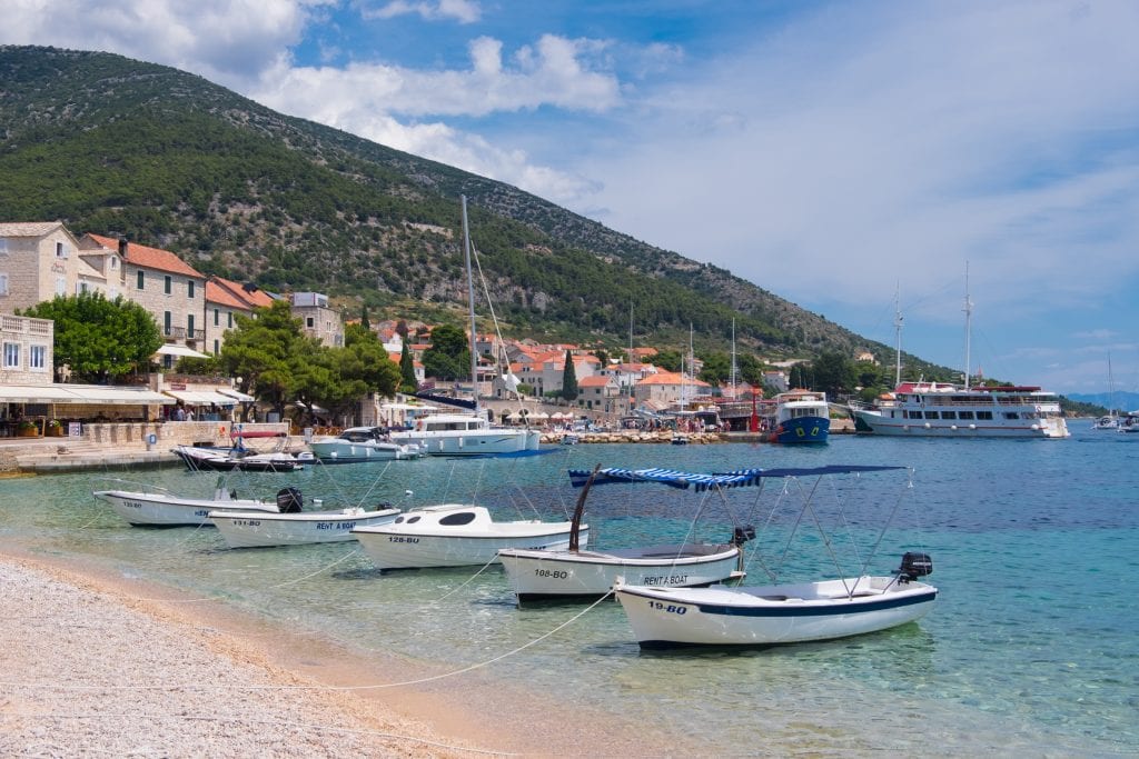 A pebble beach with clear light blue water and several small boats anchored just off shore in Brač, Croatia. In the background you see the white stone buildings of Bol topped with orange roofs.