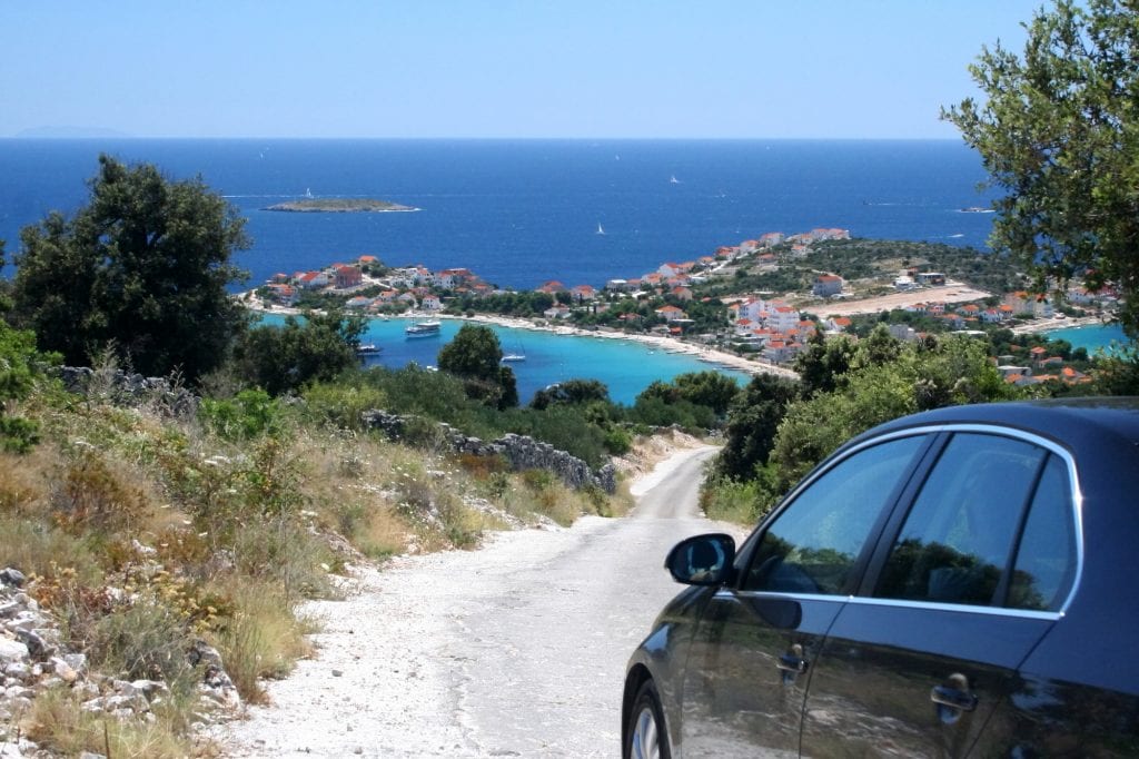 A car about to drive down a dirt road in Croatia. In the distance you see a coastal town with a bright white beach against the bright blue water.