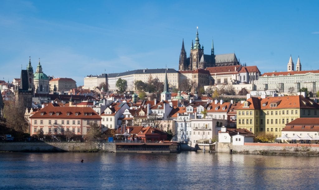 A view of Prague castle on the hill with the steeples of St. Vitus's Cathedral poking through, underneath a bright blue sky. There are lots of other colorful buildings with orange roofs, and in the foreground, the blue Vltava river.