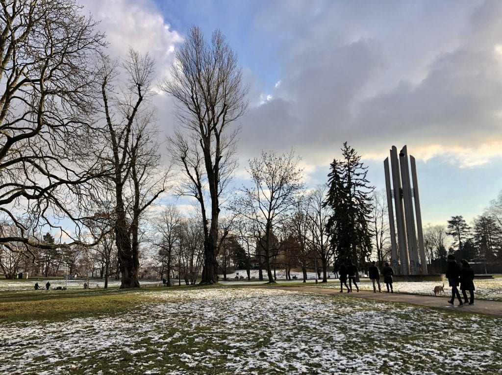 The park in Letna in Prague with grass peeking through the light snowfall, lots of leafless trees, people walking dogs on a path, and a metal sculpture of tall, thin metal cylinders.