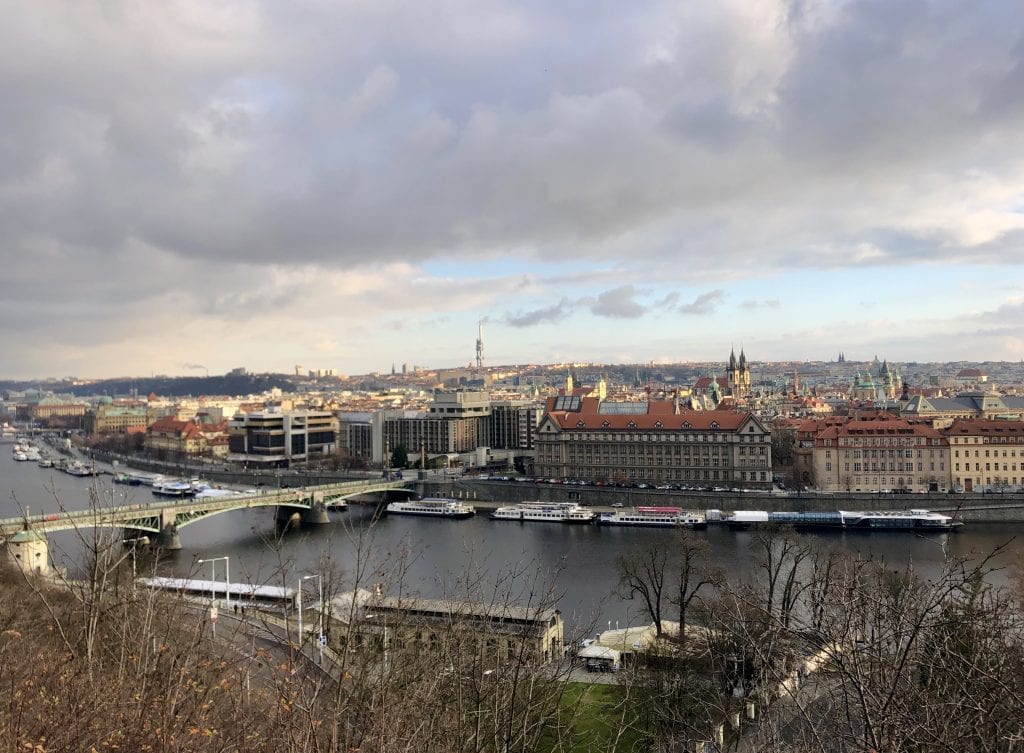 A view of Prague from a high hill in the Letna neighborhood. You see the dark gray Vltava River edged with white boats and bisected by a green bridge. Also in the background are buildings with church steeples peeking through the skyline, and the gray tube-shaped TV Tower far in the background.