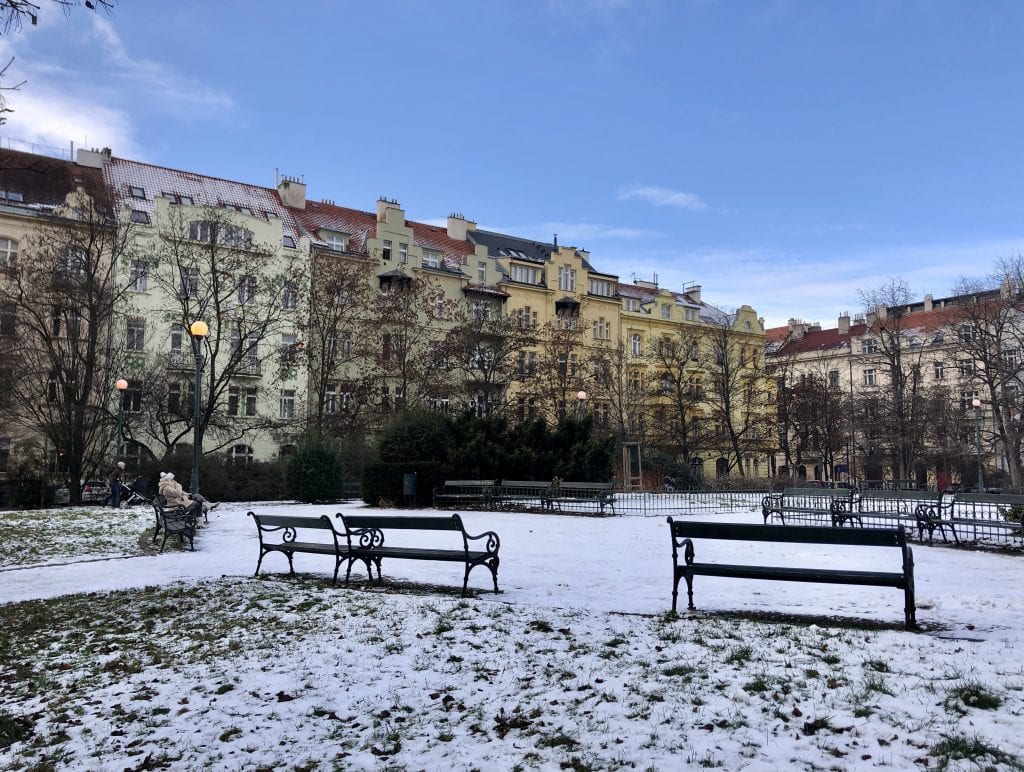 A park in Prague with light snowfall on the ground, grass peeking through, wooden benches (one with a woman sitting on it in a tan winter coat), and in the background, beige, taupe, and yellow buildings underneath a bright blue sky.