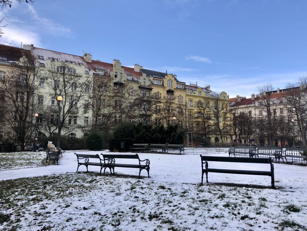 A park in Prague with light snowfall on the ground, grass peeking through, wooden benches (one with a woman sitting on it in a tan winter coat), and in the background, beige, taupe, and yellow buildings underneath a bright blue sky.