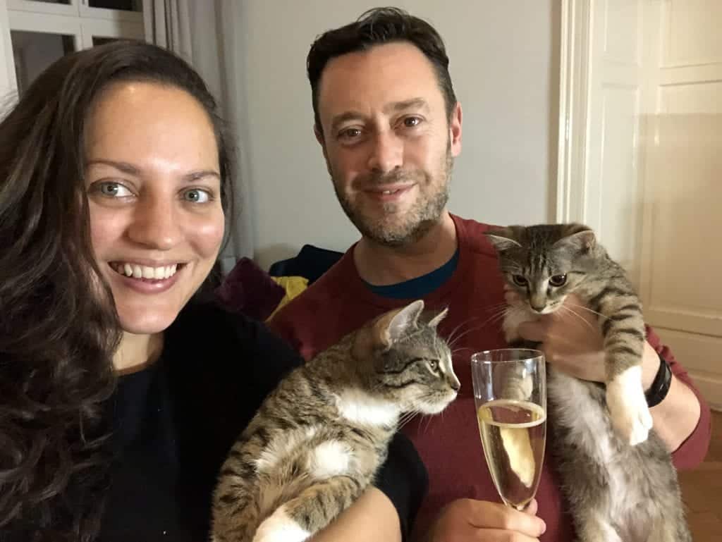 Kate and Charlie take a selfie together, each holding one of the cats. Charlie holds a glass of champagne in one hand. Both cats are looking at the champagne with curiosity.