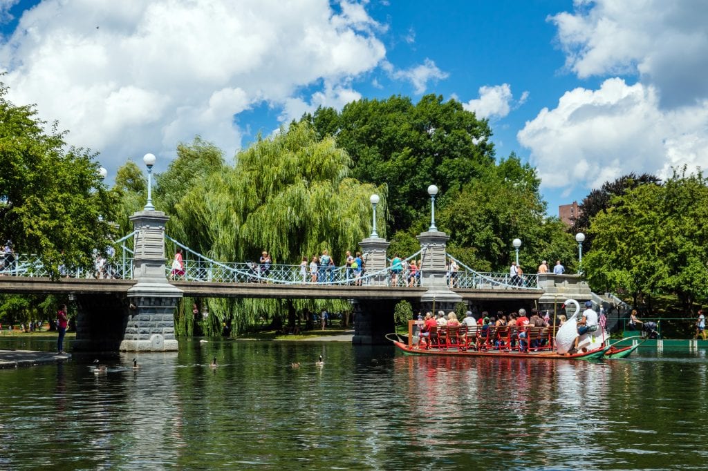 A small suspension bridge over a pond in the Boston Public Garden, a swan boat with a wooden swan in the front and people sitting on red benches, floating down the pond.