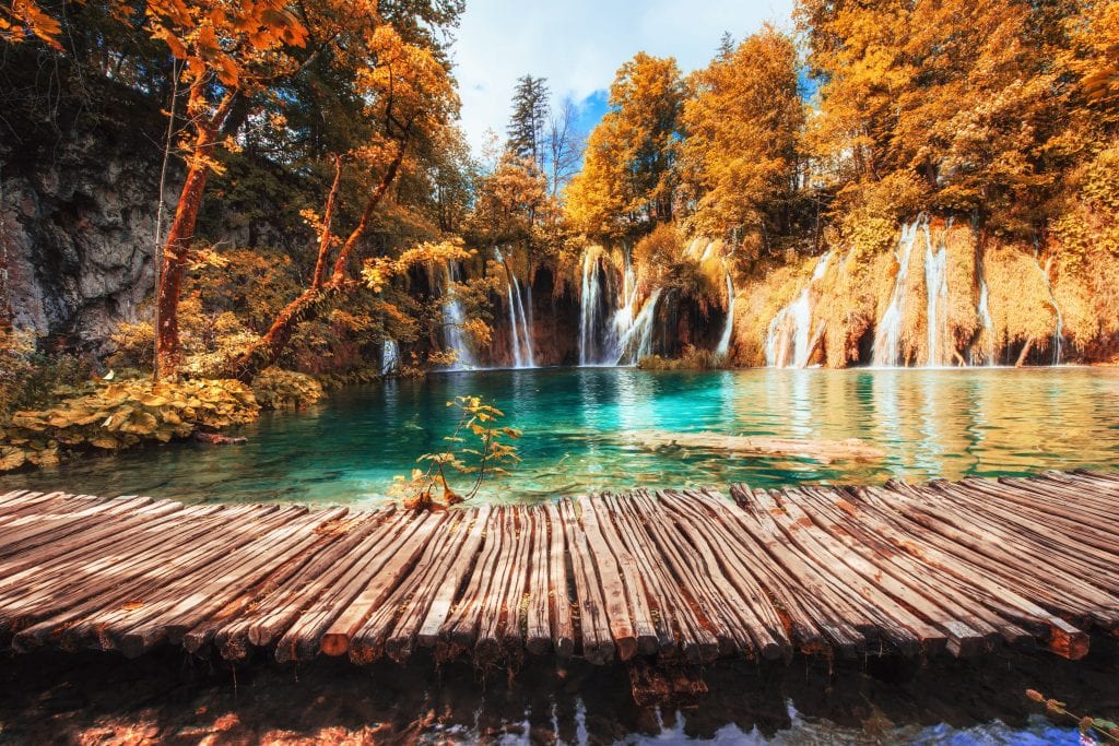 A wooden walkway on the clear teal Plitvice Lakes, surrounded by orange and red trees, waterfalls flowing through the vegetation.