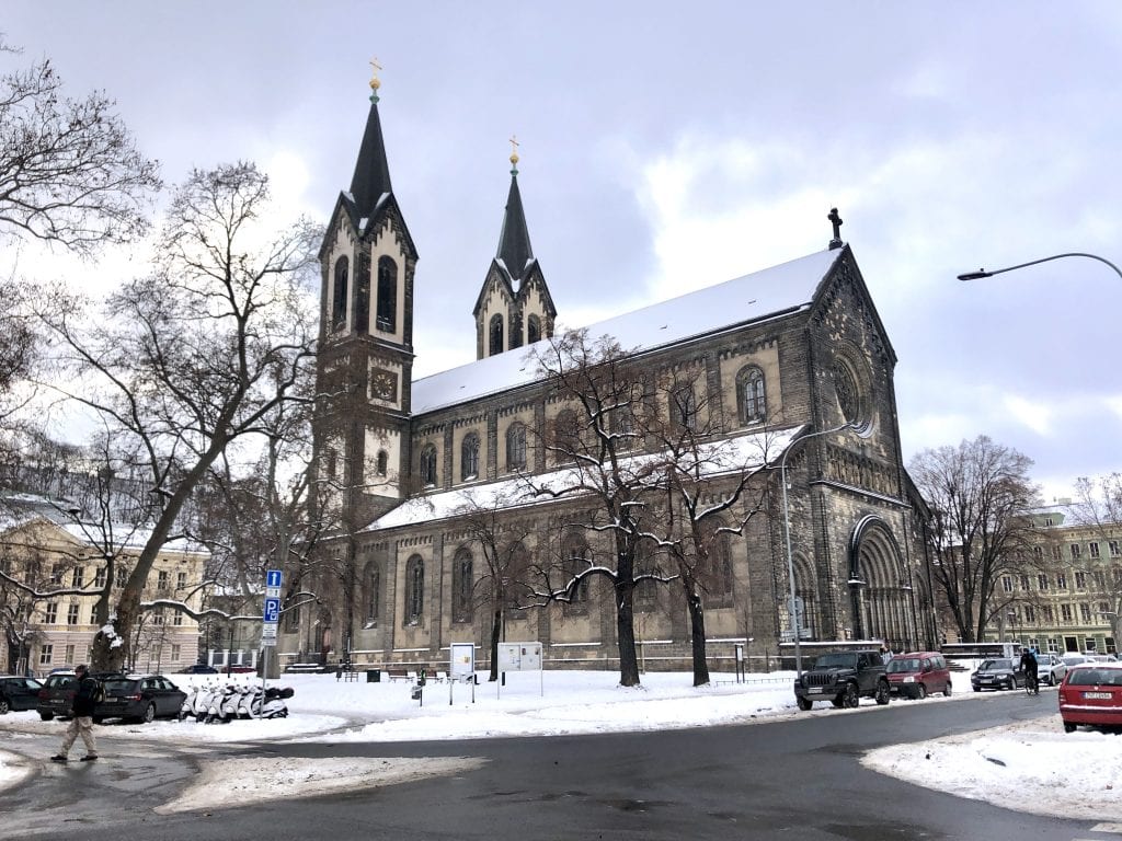 A snow-covered church on a snowy corner in Karlín, Prague, underneath a marbled cloudy sky.