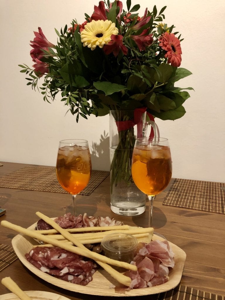 On a wooden table: in the background, a large bouquet of pink and yellow daisies with some other pink flowers and greenery. In the foreground: two bright orange aperol spritzes in wine glasses, and a wooden plate covered with various cured meats, olives, and grissini (Italian breadsticks).