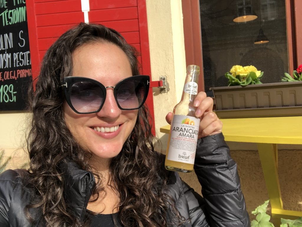Kate sits outside wearing sunglasses. She's smiling and her hair is still wet so it's crunchy, wet and very curly. She holds a bottle of Italian soda called Aranciata.