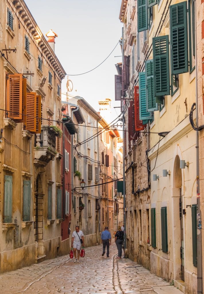 A narrow cobblestoned street in Rovinj. The buildings have green and red shutters and laundry hangs out of some buildings. It looks a lot like Italy.