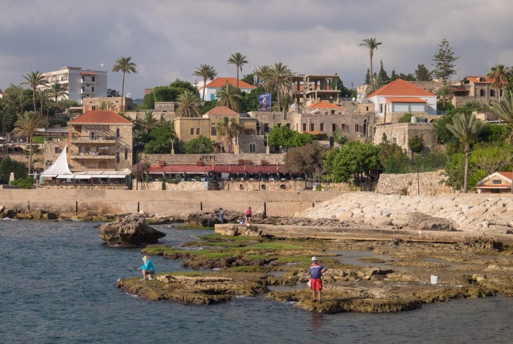 The seaside village of Byblos, Lebanon. Along the rocky edge are people standing in the water and fishing; in the background are small stone buildings on shore and lots of palm trees.