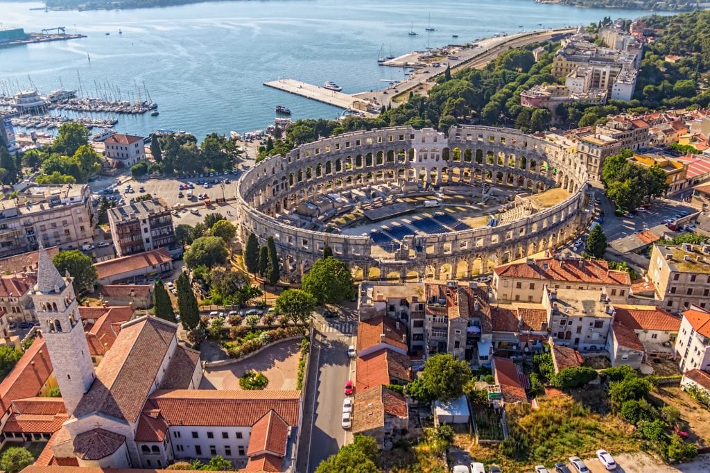 The ancient stone arena in Pula, Croatia, close to the sea. The photo is an aerial one and you see lots of terra cotta roofs and church steeples surrounding the arena.