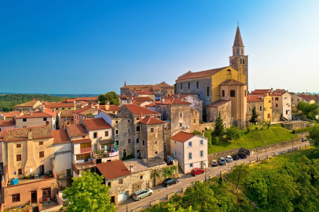 The town of Buje, Croatia, illuminated by sunshine. A small town with yellow buildings and terra cotta roofs, one church's steeple poking above the skyline, underneath a blue sky.