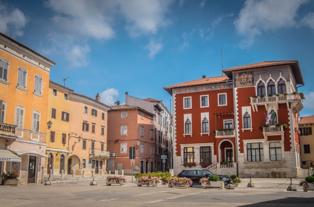 The brightly colored town of Vodnjan: yellow, peach, and blood-red buildings on an open square, underneath a bright blue sky.