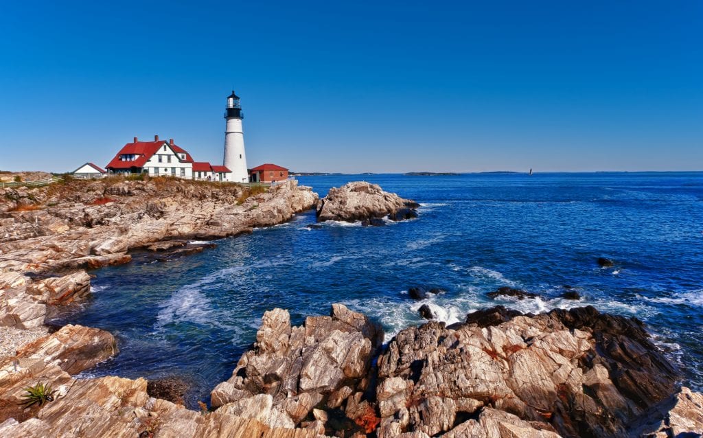 The bright white Portland Head Lighthouse, standing on a rocky outcropping jutting into the bright blue ocean.