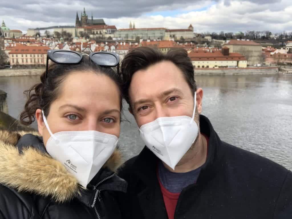 Kate and Charlie take a selfie together outside on the Charles Bridge in Prague. You can see Prague Castle rising behind them above the river. They both wear white pointy respirator face masks and smile for the camera.