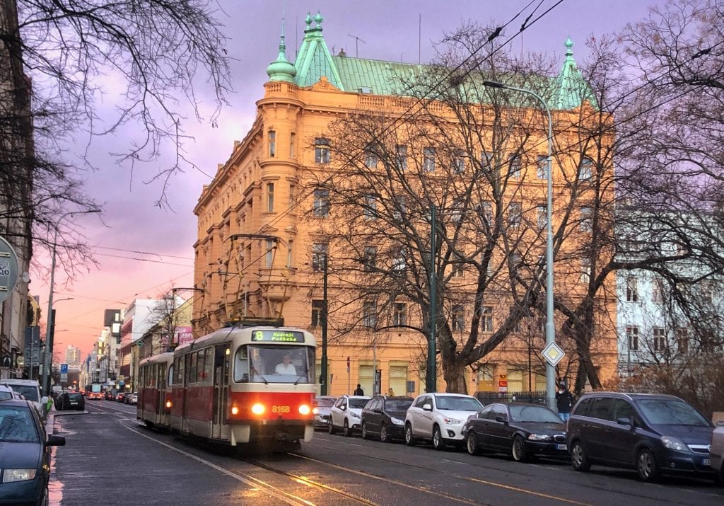 Prague in the evening: a tram comes down a street with its headlights on. In the background, a yellow building with a green roof, all set in front of an orange and purple sunset.