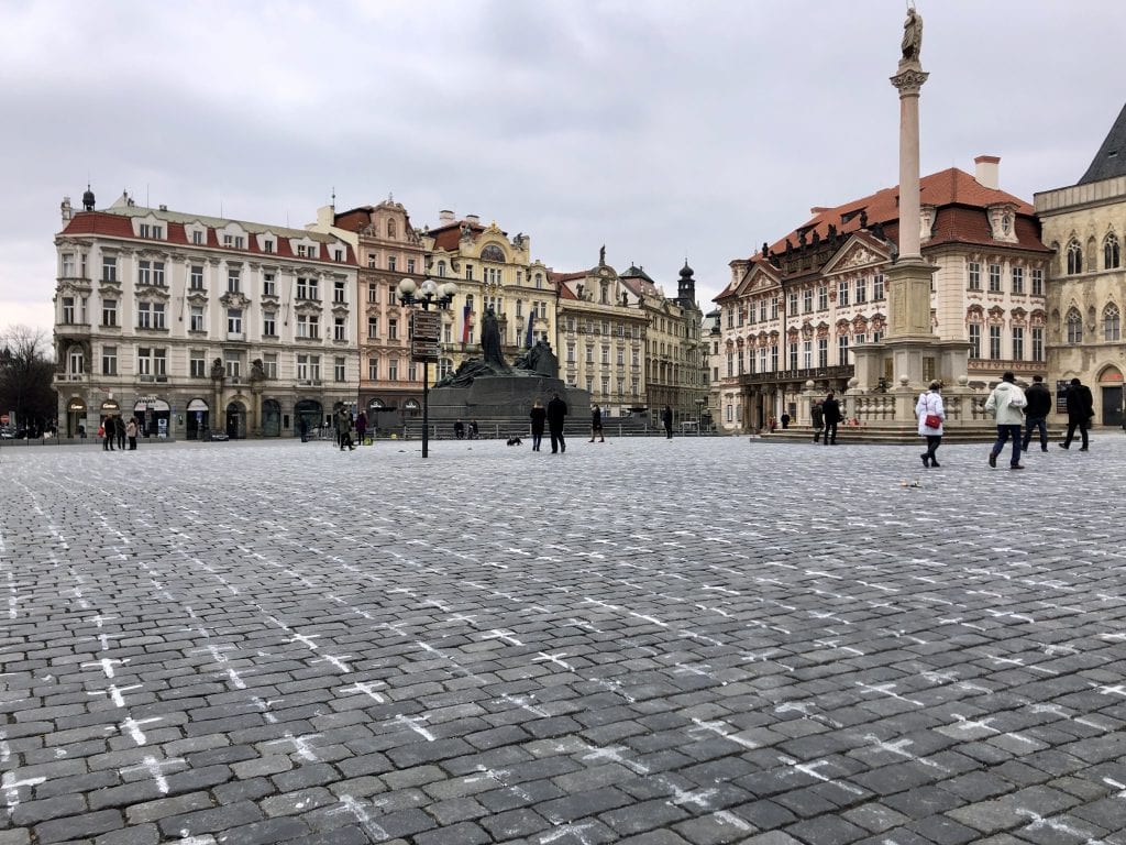 Old Town Square in Prague, edged with cream, pink and yellow crenellated buildings. In the foreground, you see the gray brick ground is covered with hundreds of white crosses, one for each Czech COVID victim.