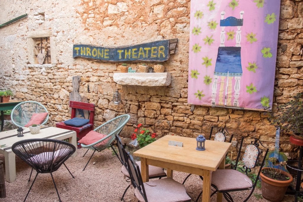A courtyard cafe. You see wooden tables with different unmatched chairs, a wooden painted sign that reads "Throne Theater", shelves with small flowerpots on them, and a painting of a chair with flowers on the background.