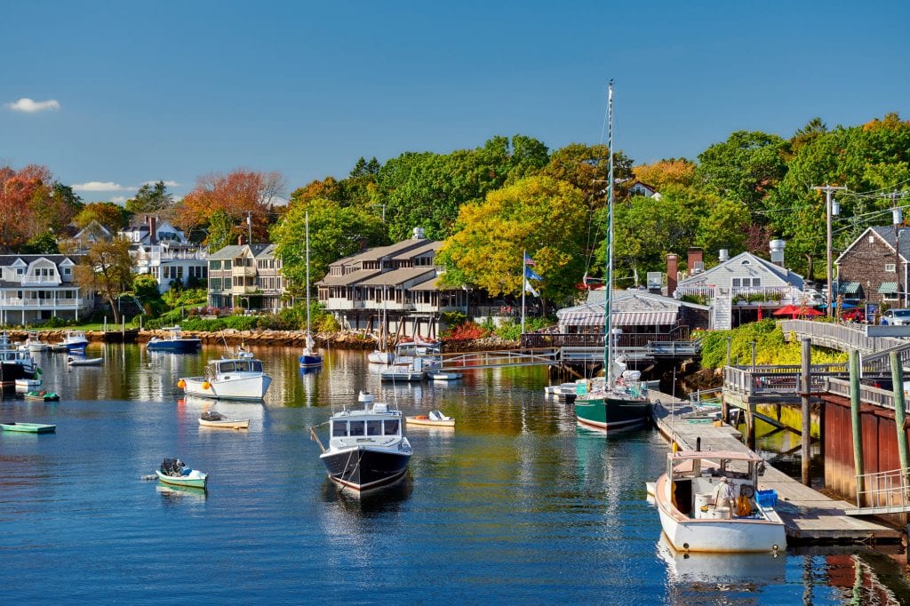 Fishing boats docked in a smooth harbor in front of waterfront homes in Ogunquit, Maine. In the background are trees just starting to turn red and yellow.