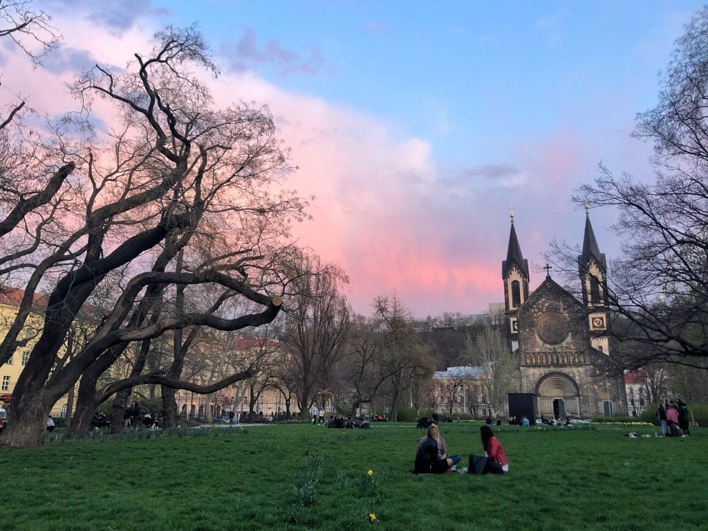 Karlinske Namesti -- my square! A green open space, like a park, in front of a church with two steeples. People are sitting in groups on the grass. The trees are still bare and the sky is a pink, blue and purple sunset.