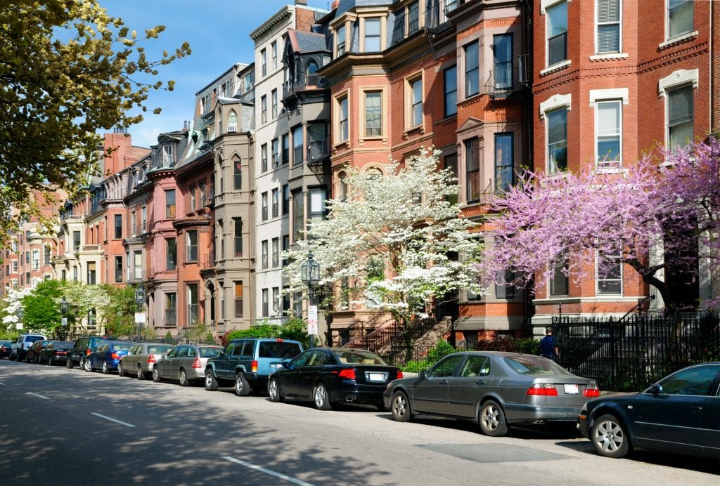 Brownstones with bay windows in Back Bay, cars parked along the sidewalk. There are several trees flowering white and pink.