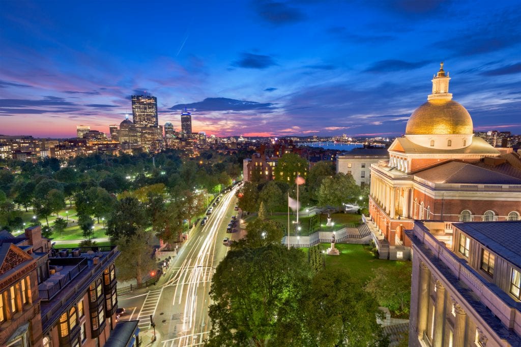 A view of Boston at night: the golden-domed state house on the right, the park of Boston Common in the foreground, and skyscrapers in the distance, underneath a sunset.