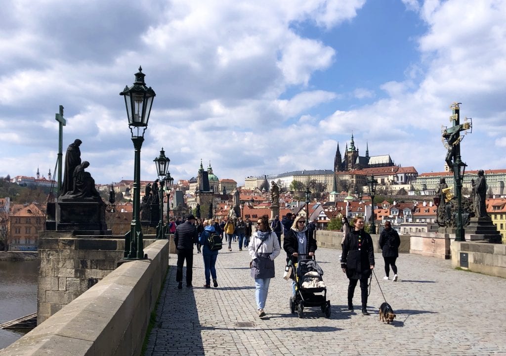 Three women, one pushing a stroller, walking along the stone Charles Bridge. You can see the steeples of the cathedral within Prague Castle poking out the background underneath a blue and white cloud-streaked sky.