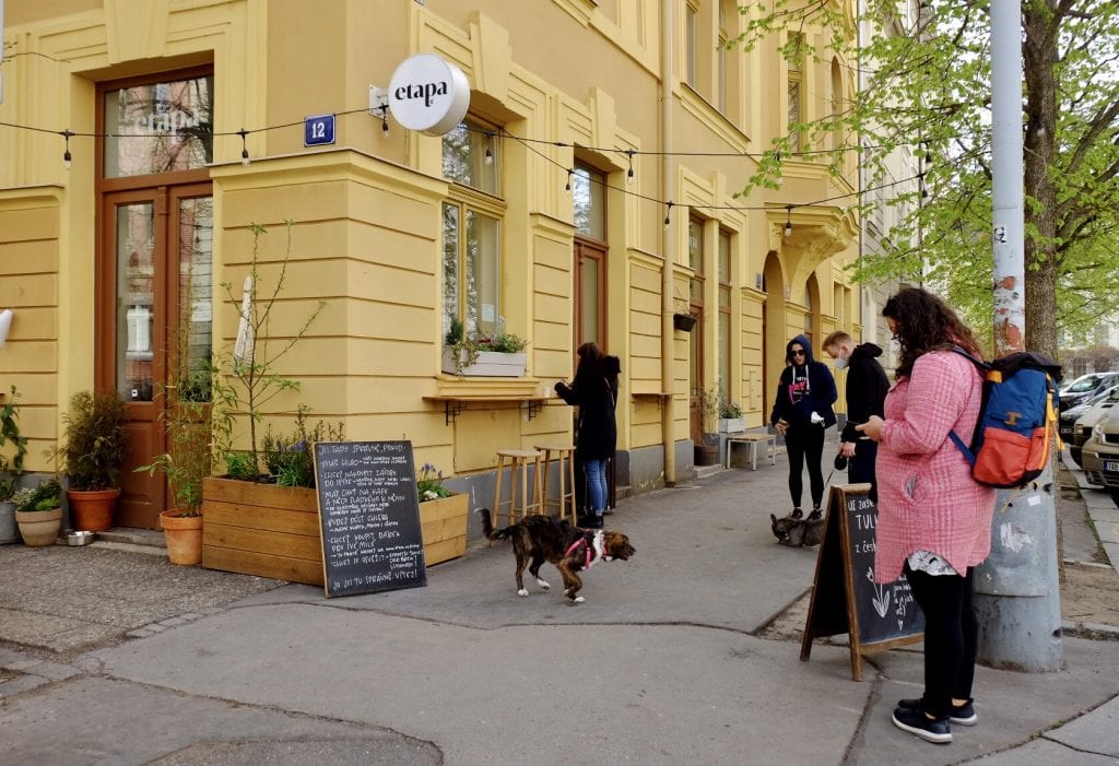 The outside of Etapa cafe, the building bright yellow, with a blackboard in front listing the specials in Czech. Four people and two dogs wait for their food outside the building.