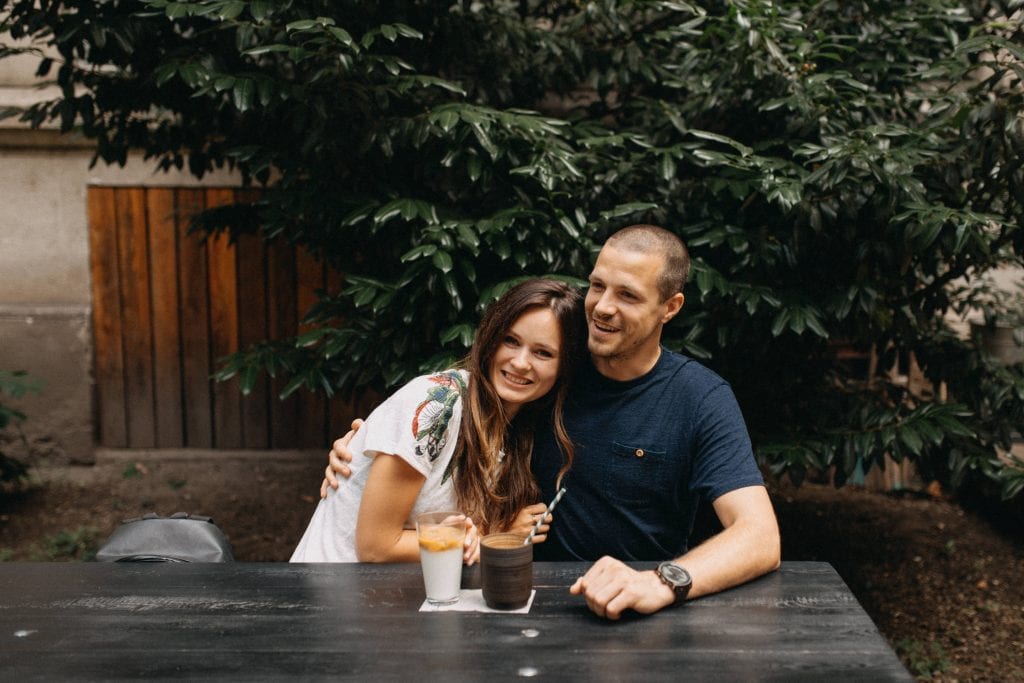 Gabi and Petr sitting at an outdoor wooden table. Gabi has long dark wavy hair and Petr has very short hair and has his arm around her. They are smiling. In front of them are two coffees.