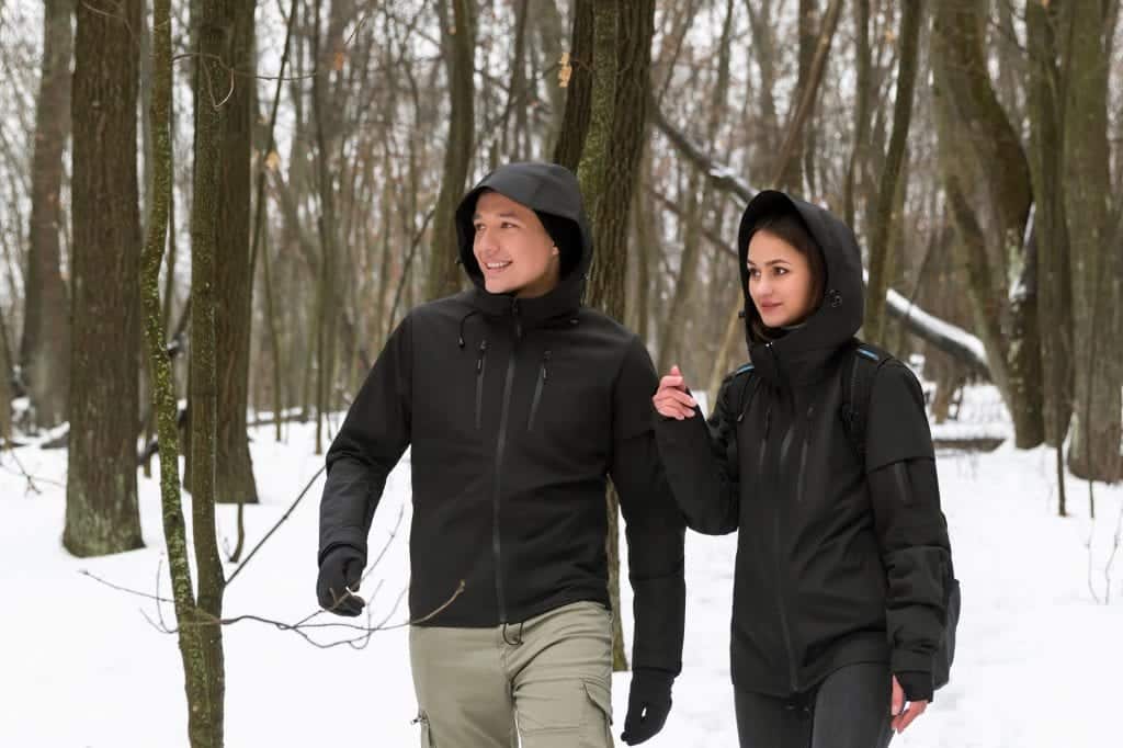 A man and a woman walking through the snowy woods, each wearing a black hooded Gamma graphene jacket.