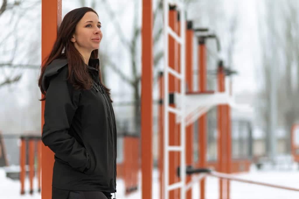 A woman standing in front of a wooden building, wearing a black gamma jacket and gazing ahead.