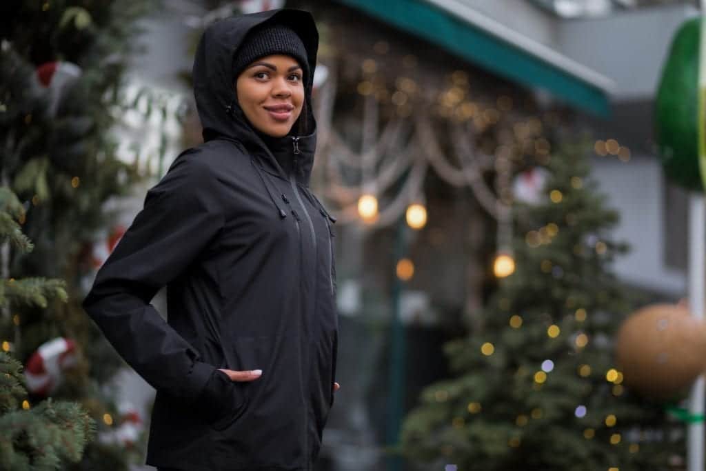 A woman wearing a black Gamma jacket with the hood up, grinning. Behind her is a Christmas tree and some twinkling lights.