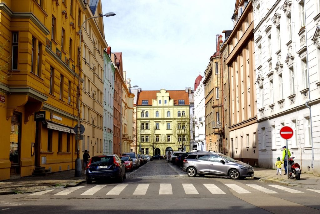 A city block in Prague: a pale yellow building in the background, crenellated with lots of angles, surrounded by a street with tan, white, and pale blue buildings, cars parked in front of each side.