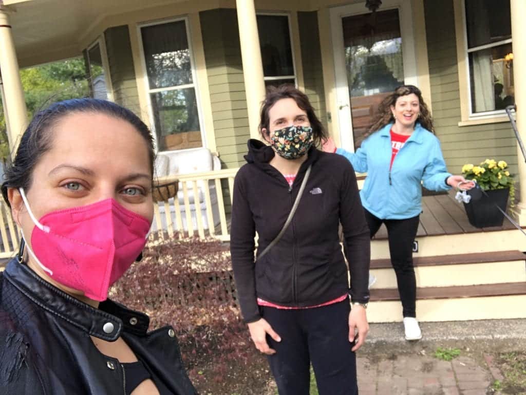 Kate and her two friends, Lisa and Alexa, standing apart from each other in front of a house. Kate wears a pink face mask, Lisa wears a flower patterned face mask, and Alexa wears no face mask but holds up her arms with a grin.