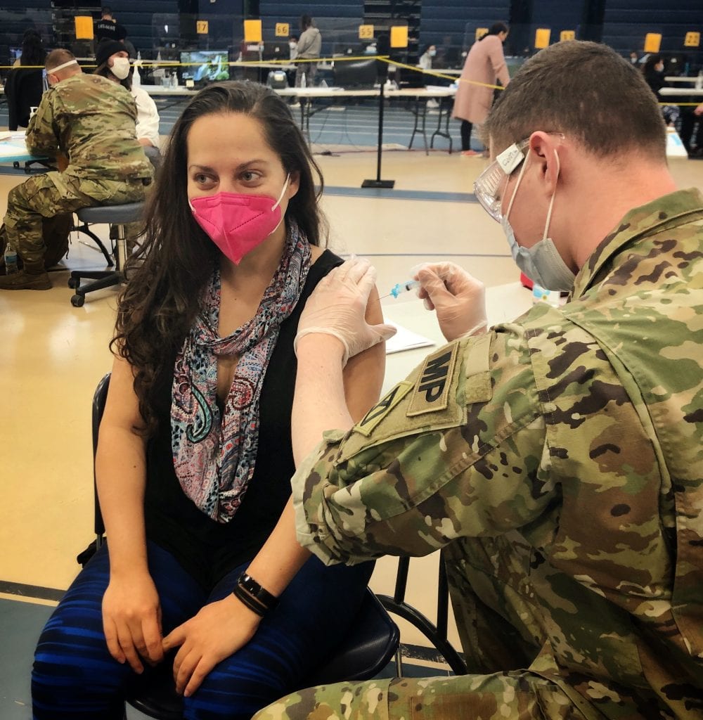 Kate sits in a chair, wearing a bright pink face mask, a black tank top, and a patterned scarf. A National Guardsman in army fatigues wearing a mask, goggles, and gloves puts the needle in her arm for the vaccine.