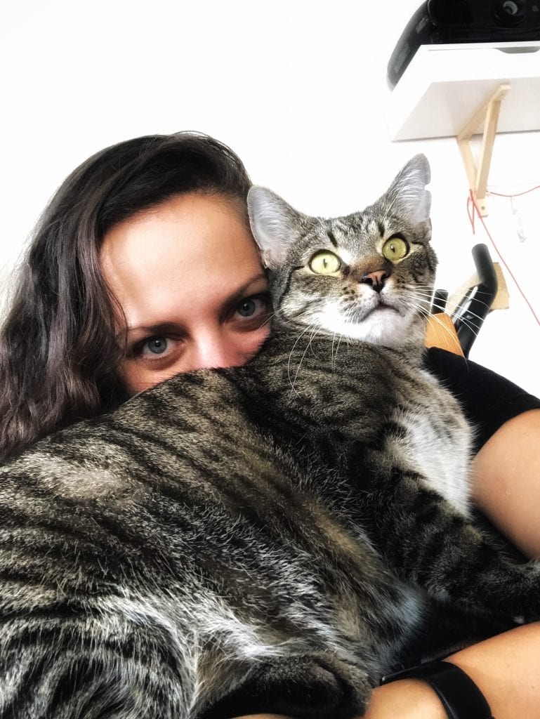 Kate holds Baliček the gray tabby cat in front of her face; you can only see her eyes peeking out from above his fur. Baliček stares straight ahead.