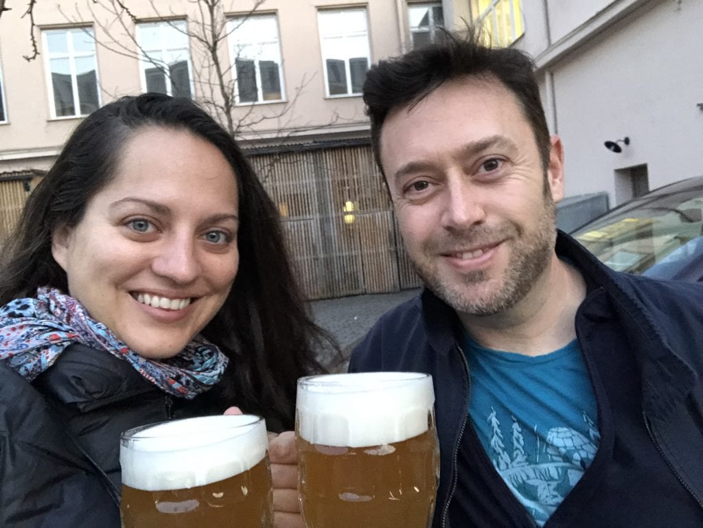 Kate and Charlie with big smiles, both wearing black coats, standing outside, each holding a pint of beer with a few inches of foam on top.