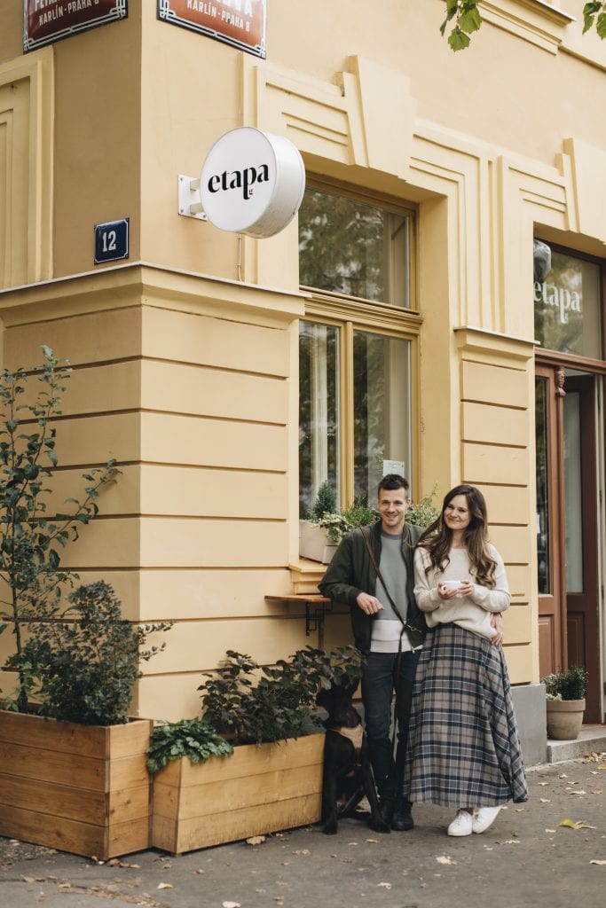 Peter and Gabi, a couple in their thirties, standing outside the cafe Etapa, which is a bright yellow building. Petr has short dark hair and wears a dark gray jacket and jeans, and Gabi has long dark wavy hair and wears a beige sweater and long gray plaid skirt.