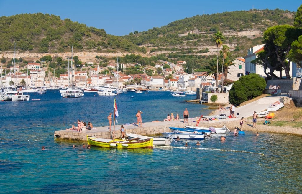 A pier jutting into the bright teal sea in Vis town, several people swimming in the water or sunning themselves. The town of Vis and several sailboats in the background.