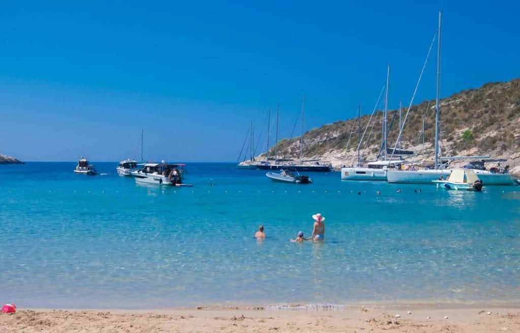 A sandy beach with calm, bright blue water. A man, woman and child playing in the water. Behind them, speedboats and sailboats.