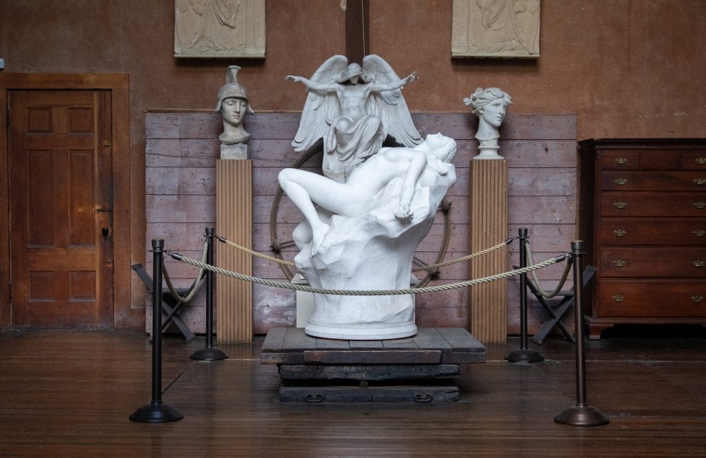 The marble statue of a naked woman lying on a rock, leaning back in ecstasy. Behind her is a marble sculpture of a super muscle angel, his arms held up as if to quiet her.