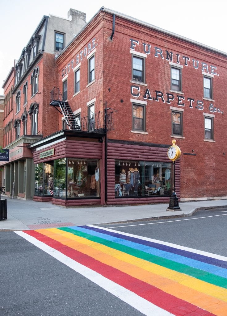 A large red brick building that used to be a furniture factory, with a boutique on the ground floor. In the foreground, a crosswalk painted rainbow colors.