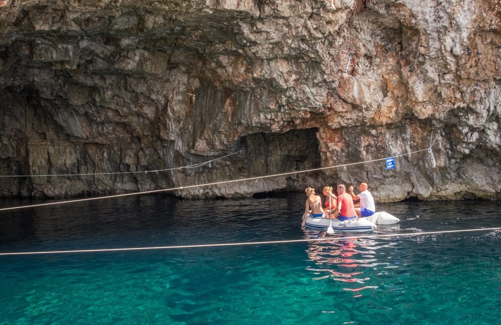 Four adults on a small inflatable boat, one paddling an oar. They are paddling into a rocky cave on top of greenish blue water.